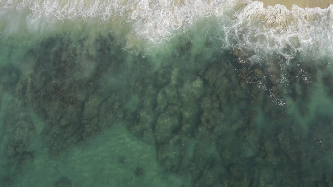 Overhead coastal view in Baja California Sur, Mexico where white wave foam spreads across the top and shadowed submerged rocks show through the green water, with no visible people.