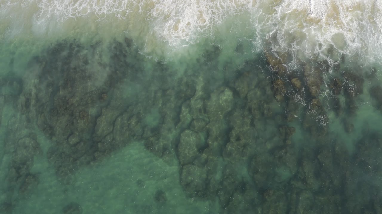 Aerial frame from Baja California Sur, Mexico with the breaking surf pushed lower into the image and a broad field of green water revealing uneven submerged rocks, with no visible people.