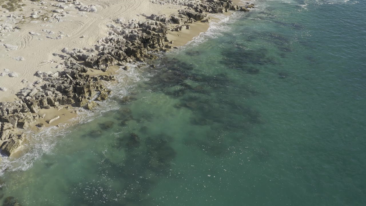 Aerial shoreline frame in Baja California Sur, Mexico, showing jagged rocks, pale beach sand, and transparent shallow water, no visible people.