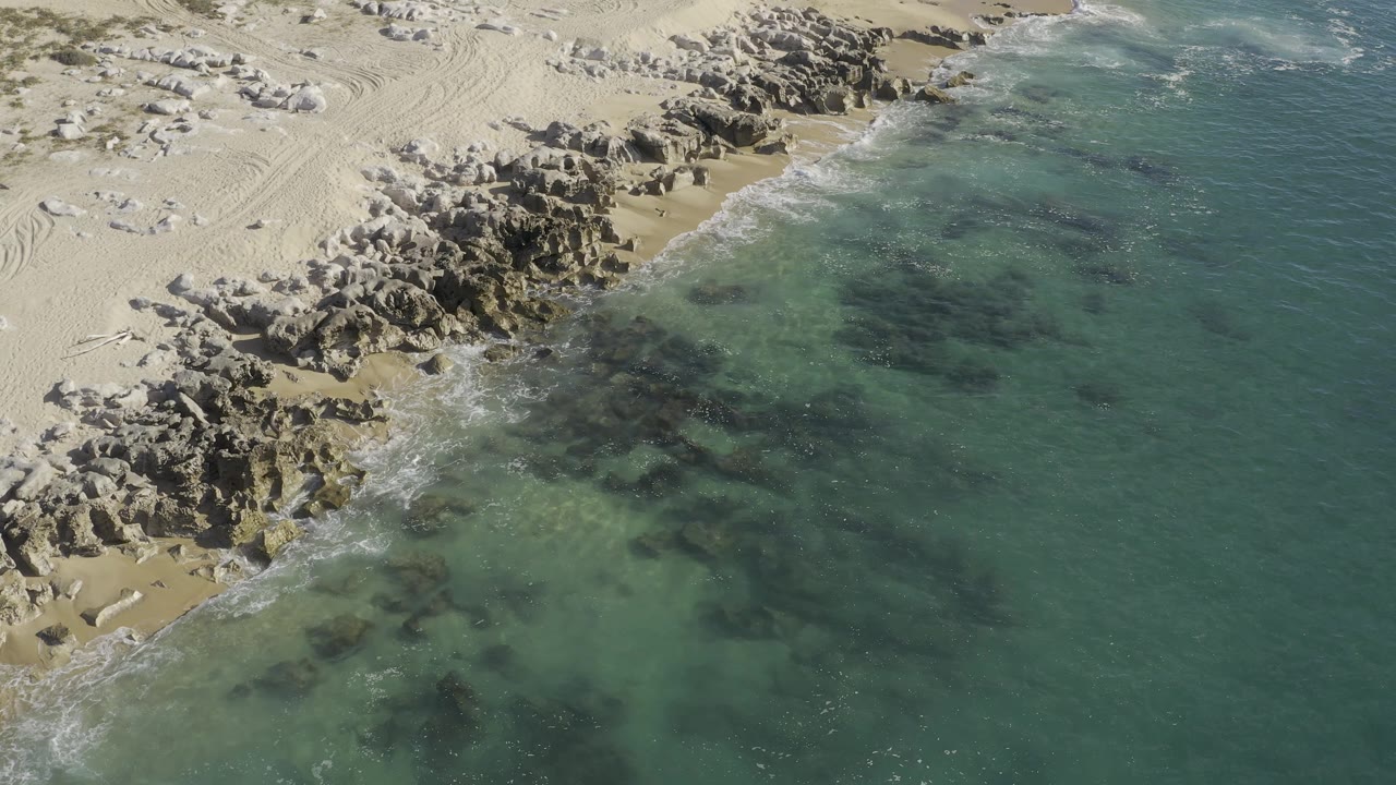 Aerial view over Baja California Sur, Mexico, with clustered shoreline rocks at left and clear coastal water spreading across the right side, no visible people.