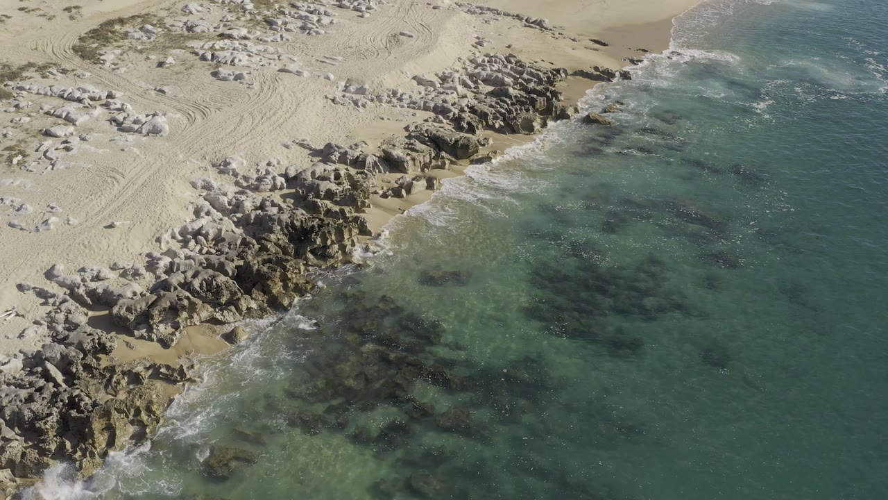 Overhead view in Baja California Sur, Mexico, showing sand tracks above rocky outcrops and clear surf wrapping the shore, no visible people.
