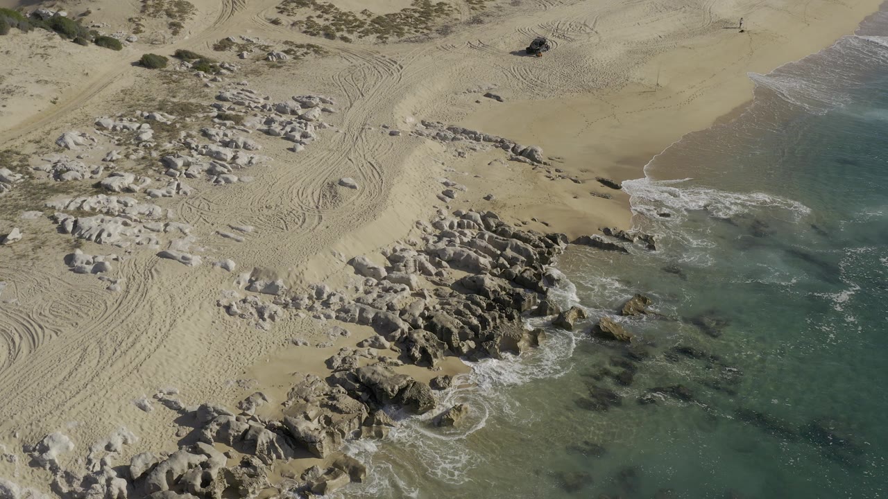 Overhead view in Baja California Sur, Mexico, showing pale sand, tire tracks, scattered rocks, turquoise surf along the right edge, and a small dark pickup with no visible people.