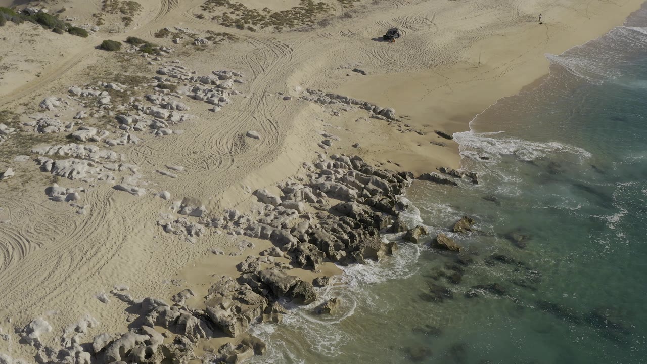 Aerial frame from Baja California Sur, Mexico, with rock clusters centered near the surf, curved vehicle tracks across the sand, a dark pickup high in frame, and no visible people.