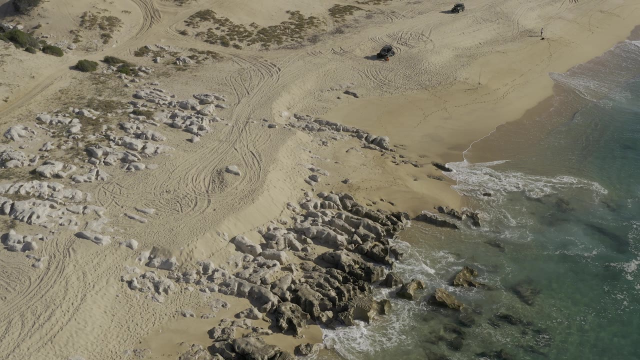 High aerial view in Baja California Sur, Mexico, where the water sits along the right side, rocks run diagonally through the lower frame, tire tracks cross open sand, and no visible people appear.