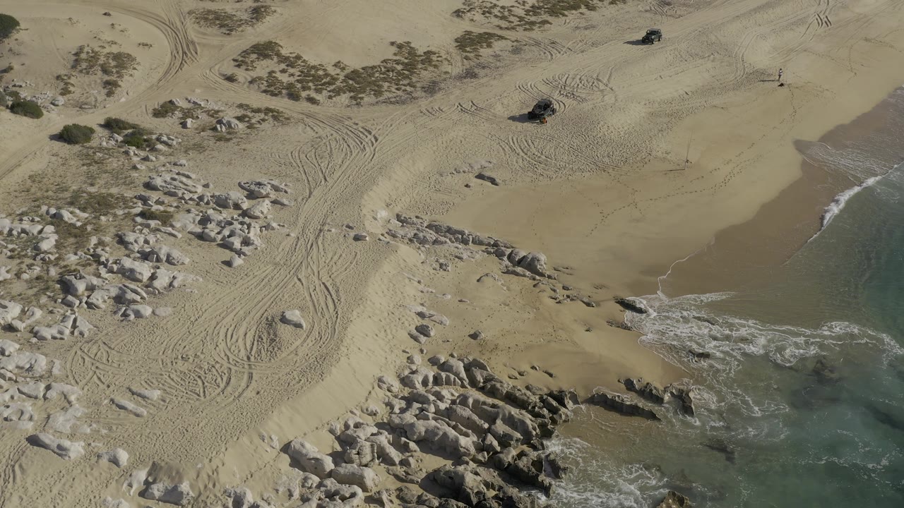 Aerial view in Baja California Sur, Mexico, showing a wider patch of smooth sand near the water, dark rocks at the lower left, looping tire tracks, a pickup near the top, and no visible people.