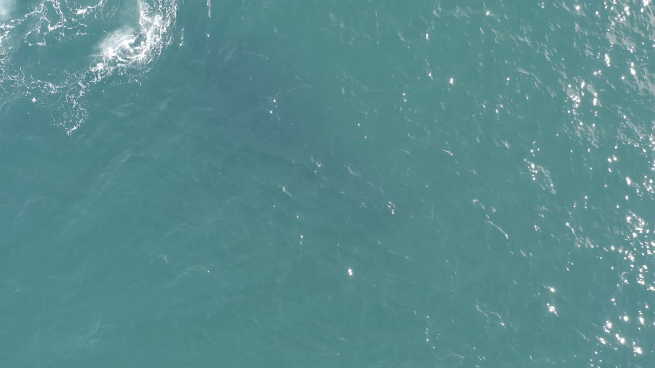 Baja California Sur ocean surface seen from above, with turquoise water, white foam activity near the upper left and top edge, and no visible people.