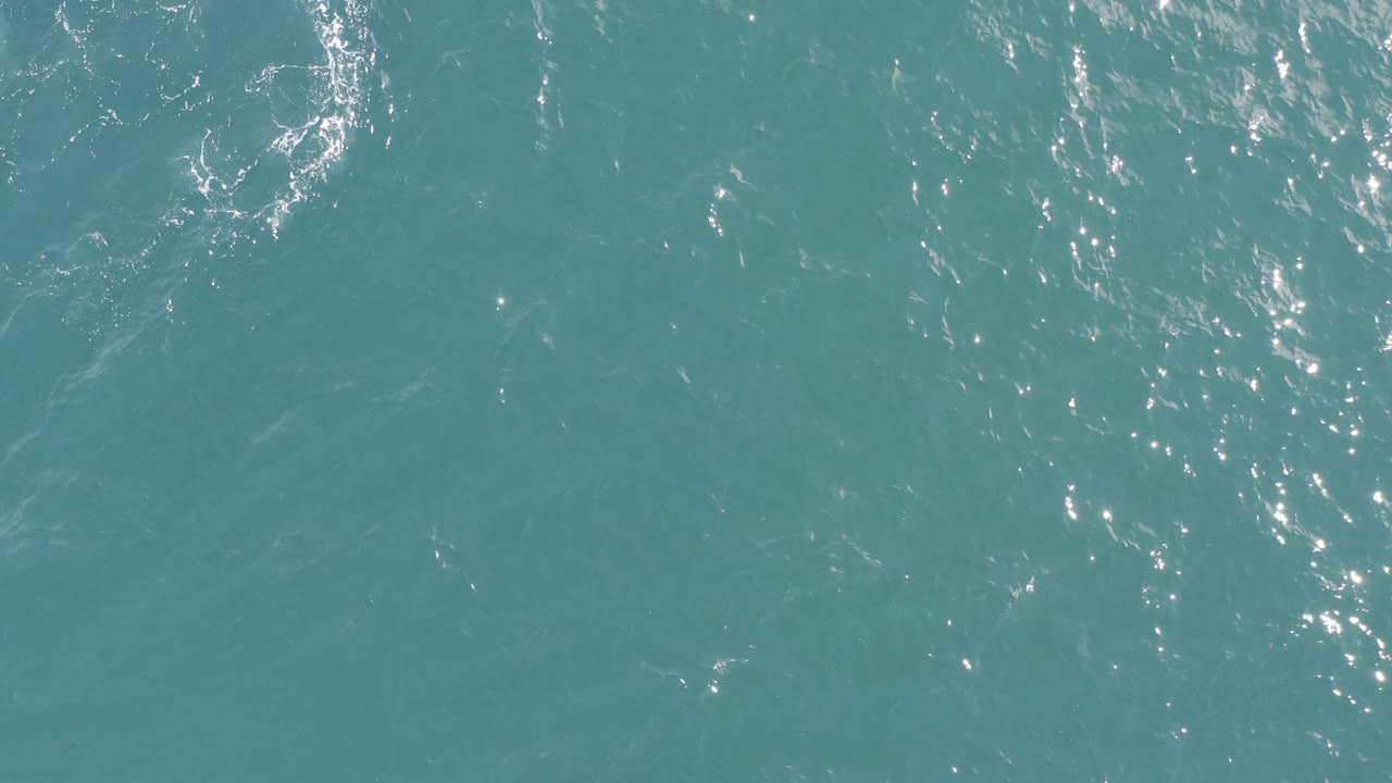 Blue-green sea water in Baja California Sur, Mexico, seen from above with scattered white flecks of light and soft diagonal current marks, no visible people.