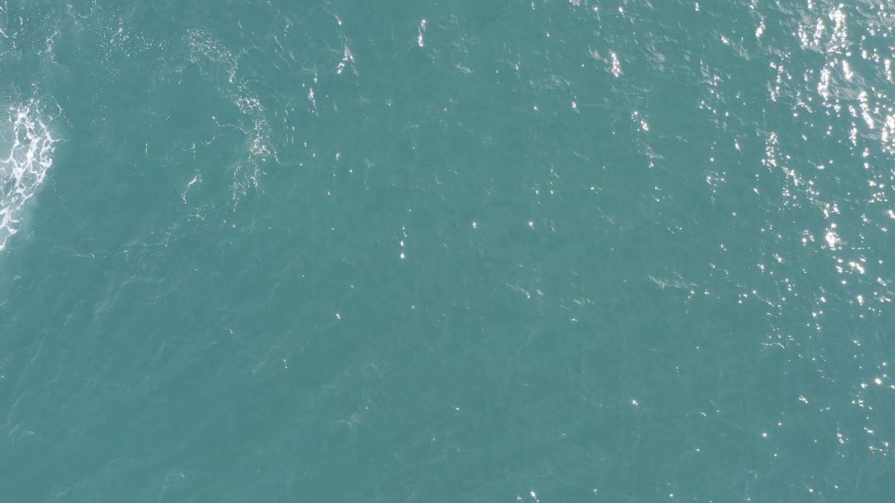 Turquoise ocean surface with sun glints and a pale circular ripple near the lower left.