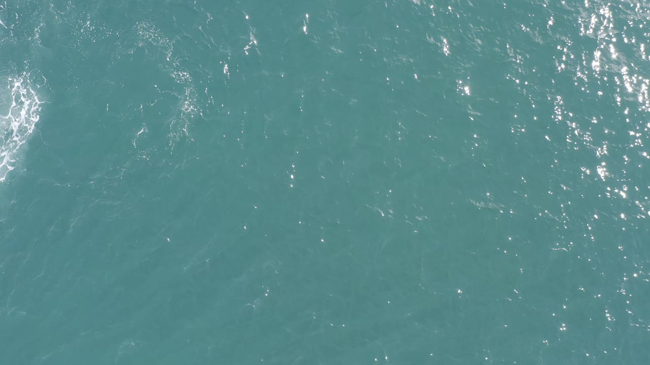 Overhead turquoise sea in Baja California Sur, Mexico, with a vertical cluster of bright sun glints and a soft pale disturbance near the center, no visible people.