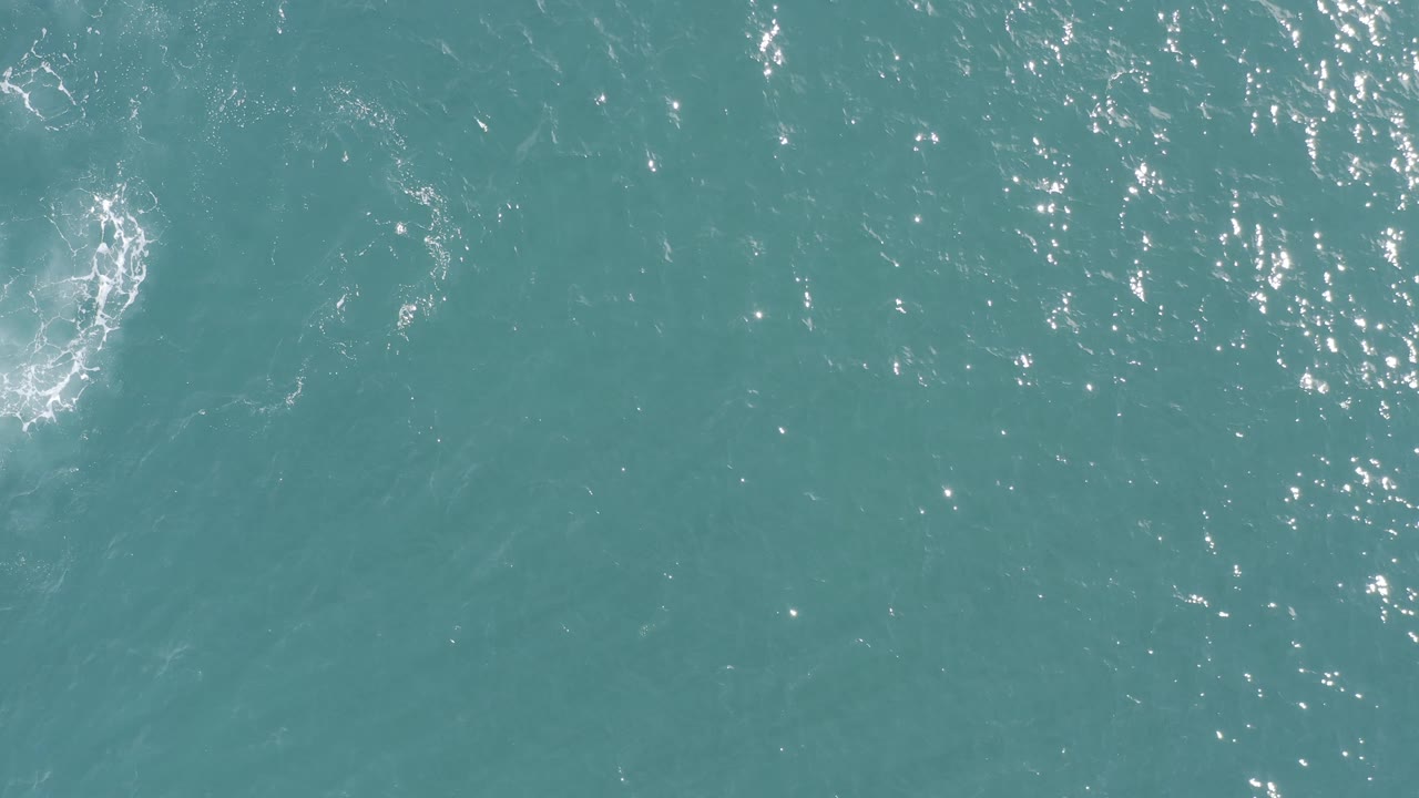 Top-down ocean surface in Baja California Sur, Mexico, showing scattered sun reflections and a compact white circular ripple in the upper right area, no visible people.