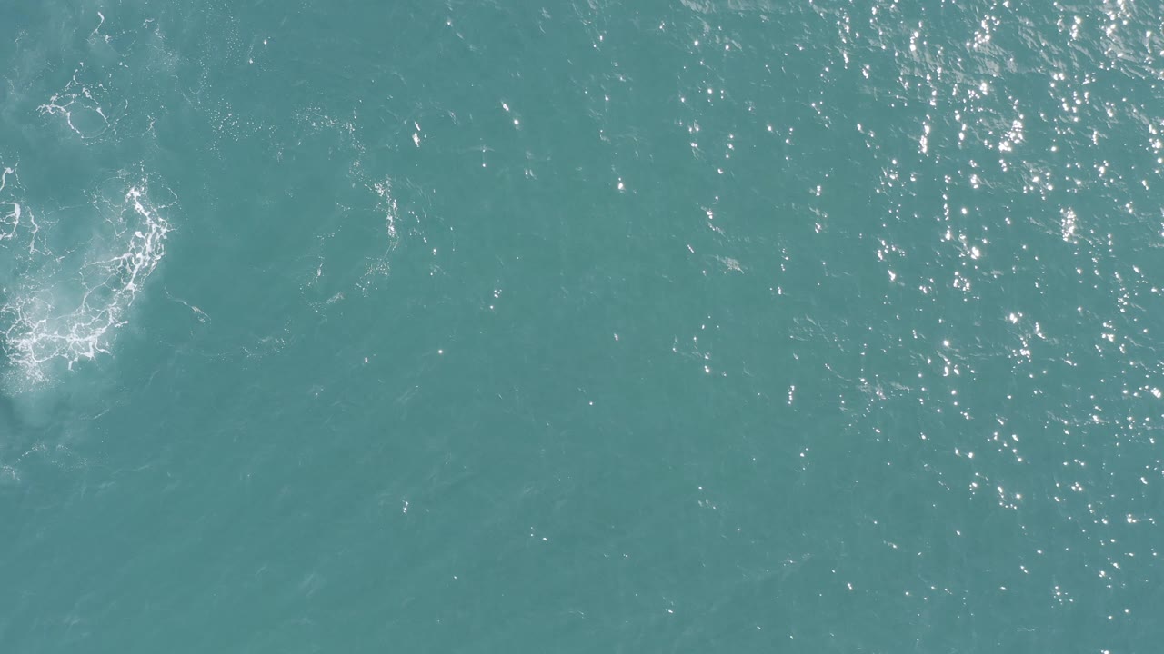 Aerial view of Baja California Sur ocean water with a pale circular foam patch along the left edge and fine sun flecks across the surface, no visible people.