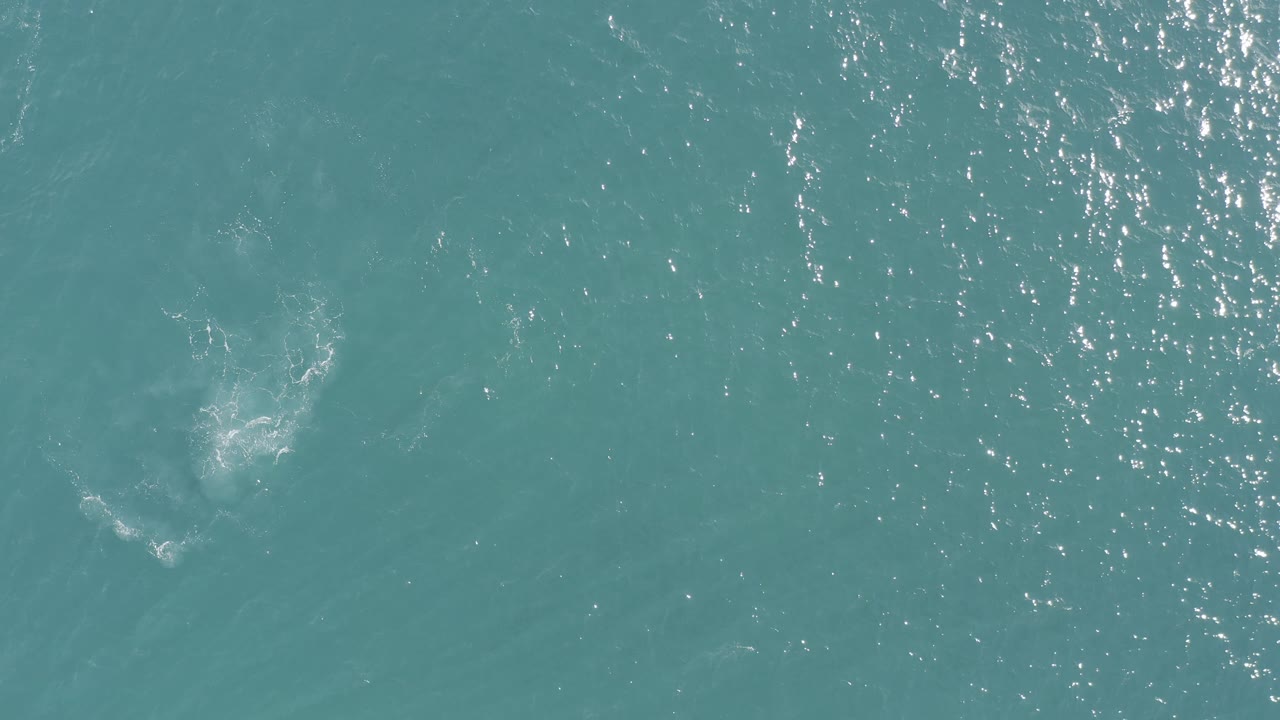 Aerial view over blue-green water in Baja California Sur, where a pale foam burst sits near the upper middle and the rest of the frame remains open water with no visible people.