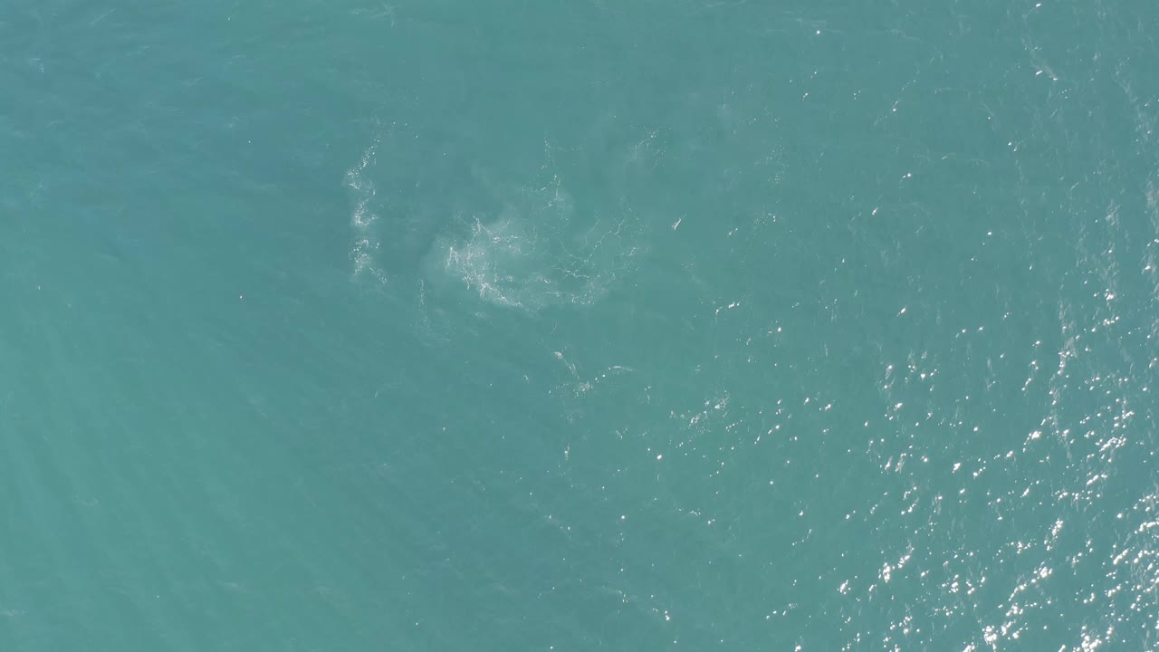 Aerial frame over blue-green water in Baja California Sur, Mexico, showing bright surface glints and a soft pale patch near the upper center, with no visible people.