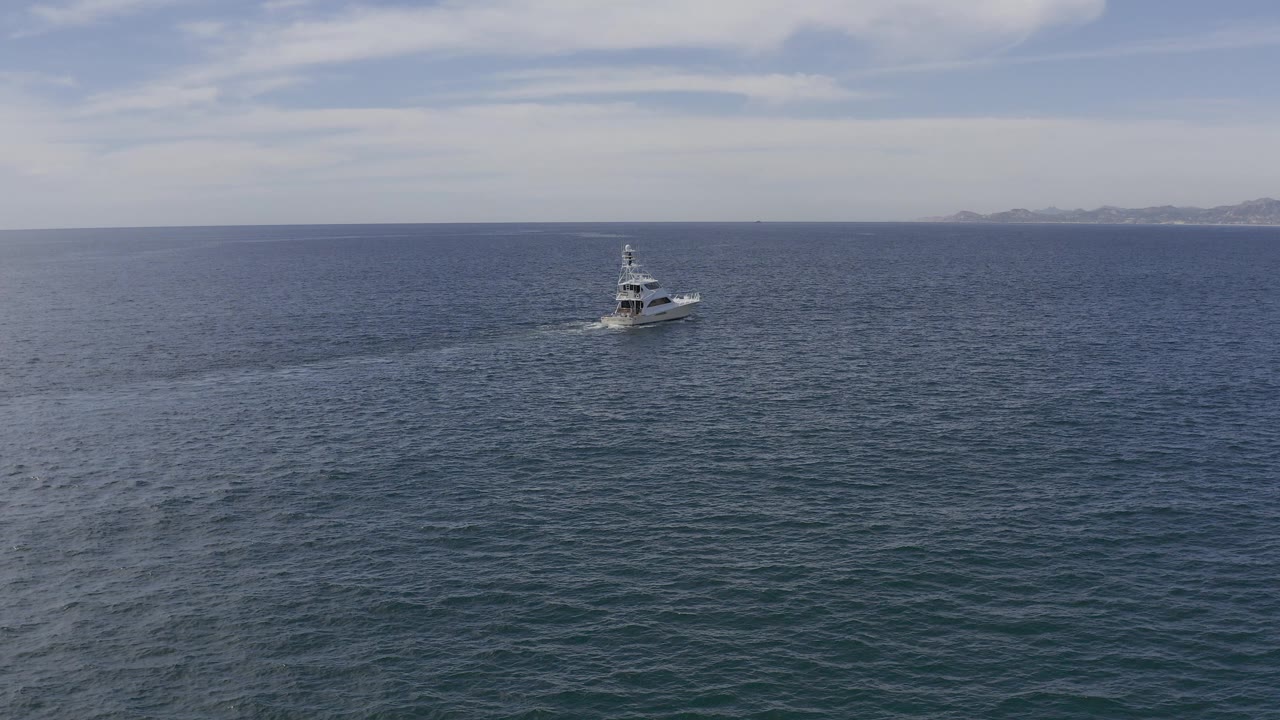 Wide aerial view in Baja California Sur, Mexico, showing a white motor yacht on open blue water with a faint wake, distant low land, pale sky, and no visible people.