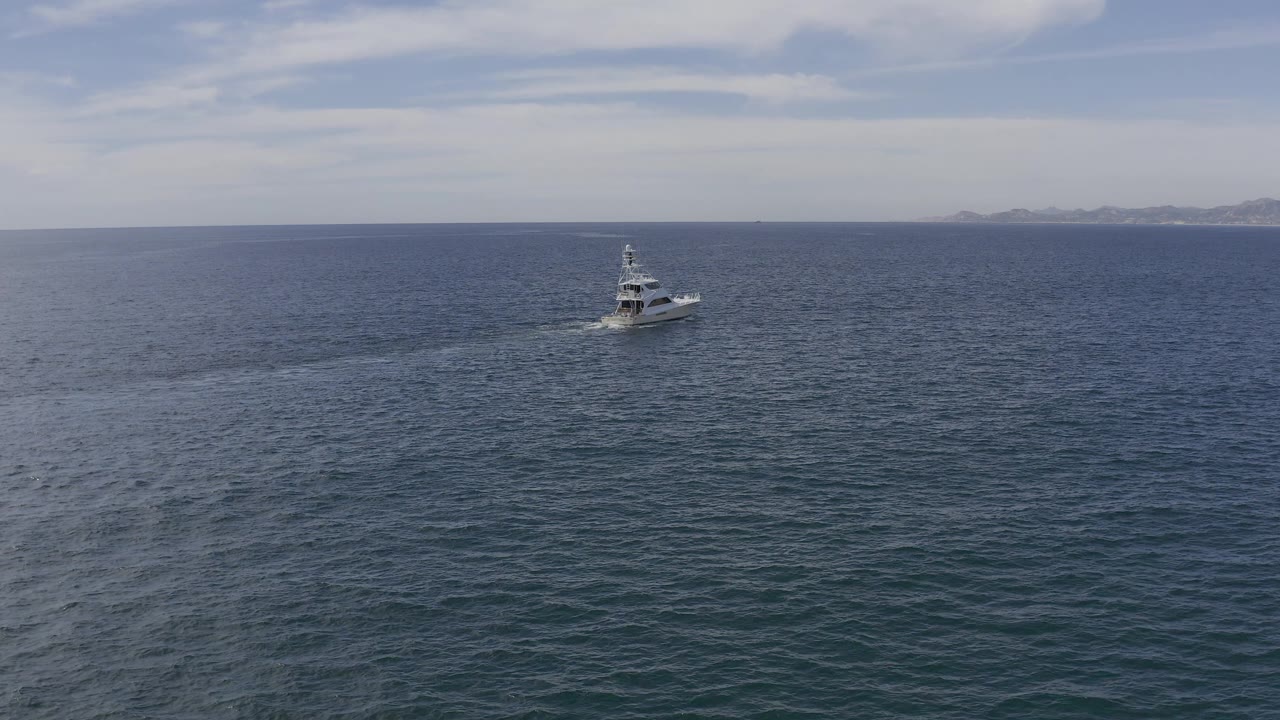 Aerial frame from Baja California Sur, Mexico, with the white yacht slightly farther across rippled blue water, a trailing wake, distant coastline, and no visible people.