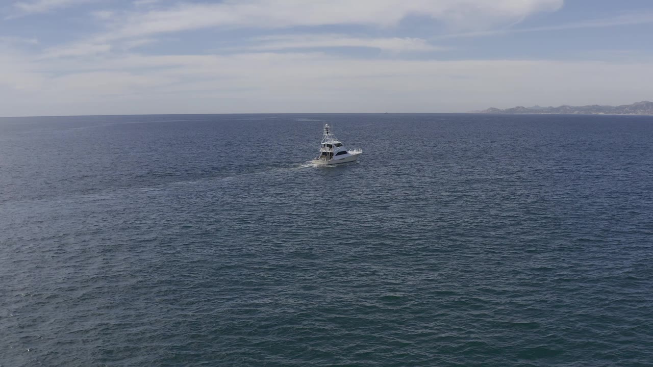 High wide view in Baja California Sur, Mexico, showing the yacht shifted across the frame on dark blue ocean, with a longer visible wake, hazy land on the horizon, and no visible people.