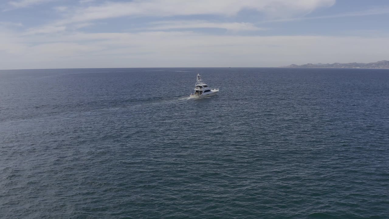 Baja California Sur aerial frame with the motor yacht small against textured blue water, a white wake behind it, muted sky above, distant land, and no visible people.