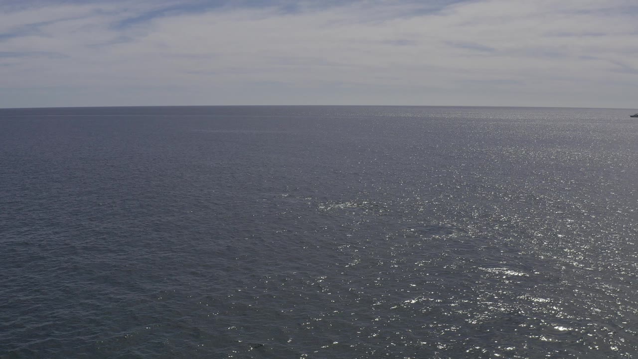 Wide aerial view over blue ocean in Baja California Sur, with a low horizon, distant land near the top of frame, sun glare on the water, and no visible people.