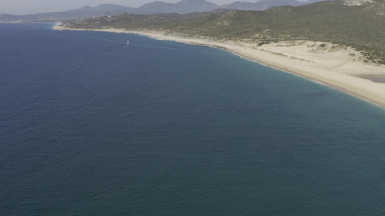 Aerial view over Baja California Sur coastline with dark blue ocean, pale beach, scrub hills, distant mountains, and no visible people at the start of the clip.