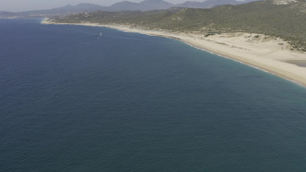 Elevated view of the Baja California Sur beach where dark blue water transitions toward lighter shallows near pale dunes, with hills and mountains in the background and no visible people.
