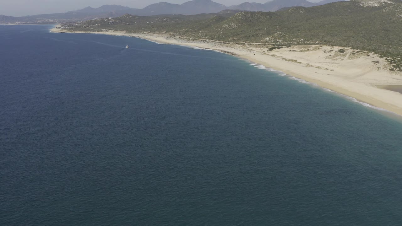 Aerial view in Baja California Sur, Mexico, with a small sailboat offshore, dark blue water across the lower frame, pale sand and dunes on the coast, and no visible people.