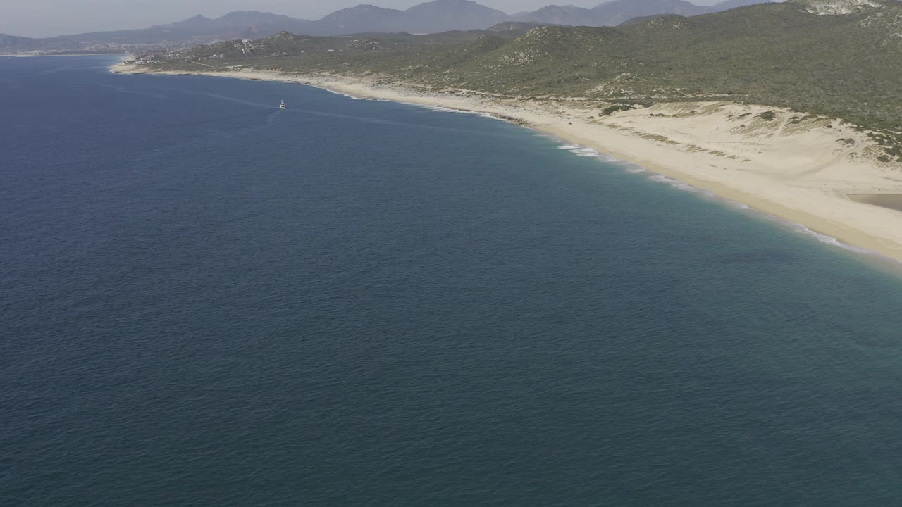 Aerial frame from Baja California Sur, Mexico, looking across broad blue ocean toward a long sandy beach, low dunes, green hills, distant mountains, and no visible people.