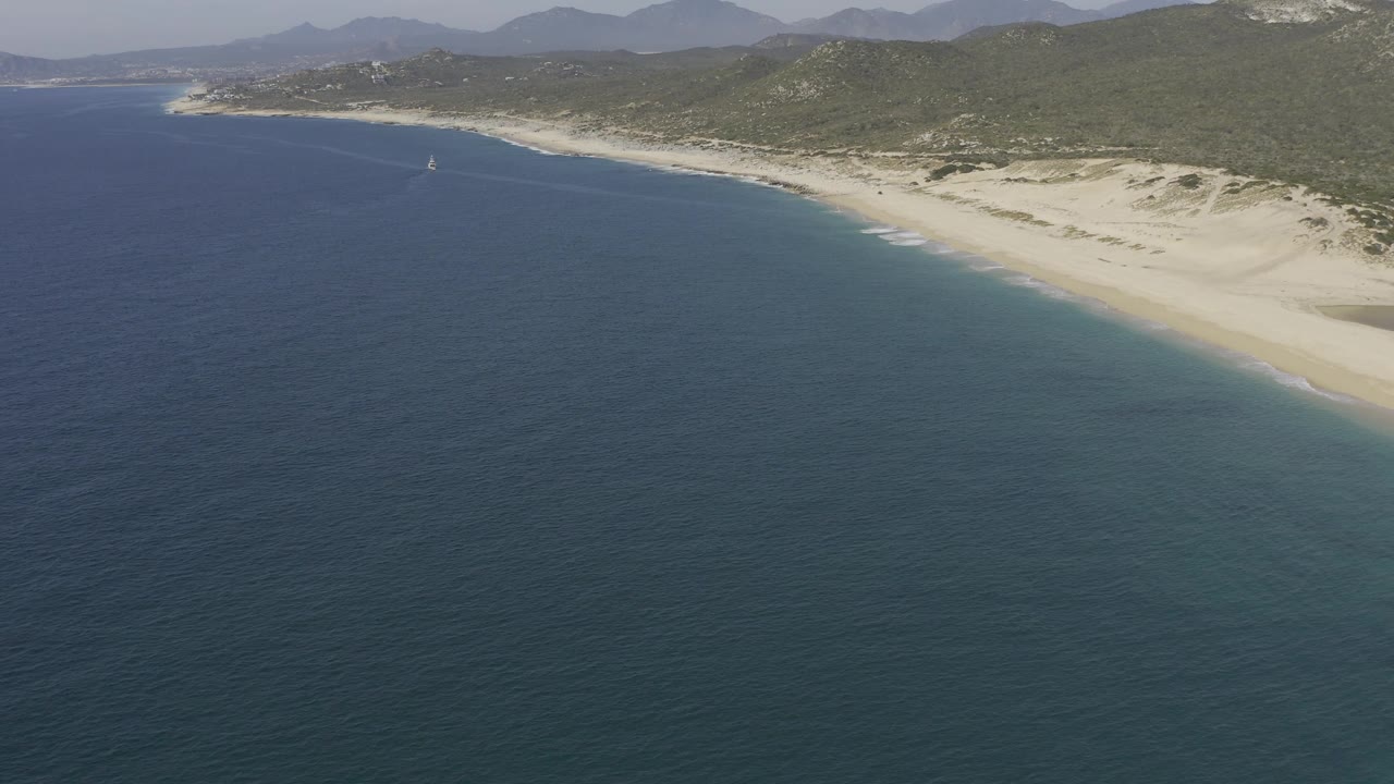 Baja California Sur aerial frame with textured blue water in front, a bright sandy coast and dunes to the right, green hills beyond the beach, and no visible people.
