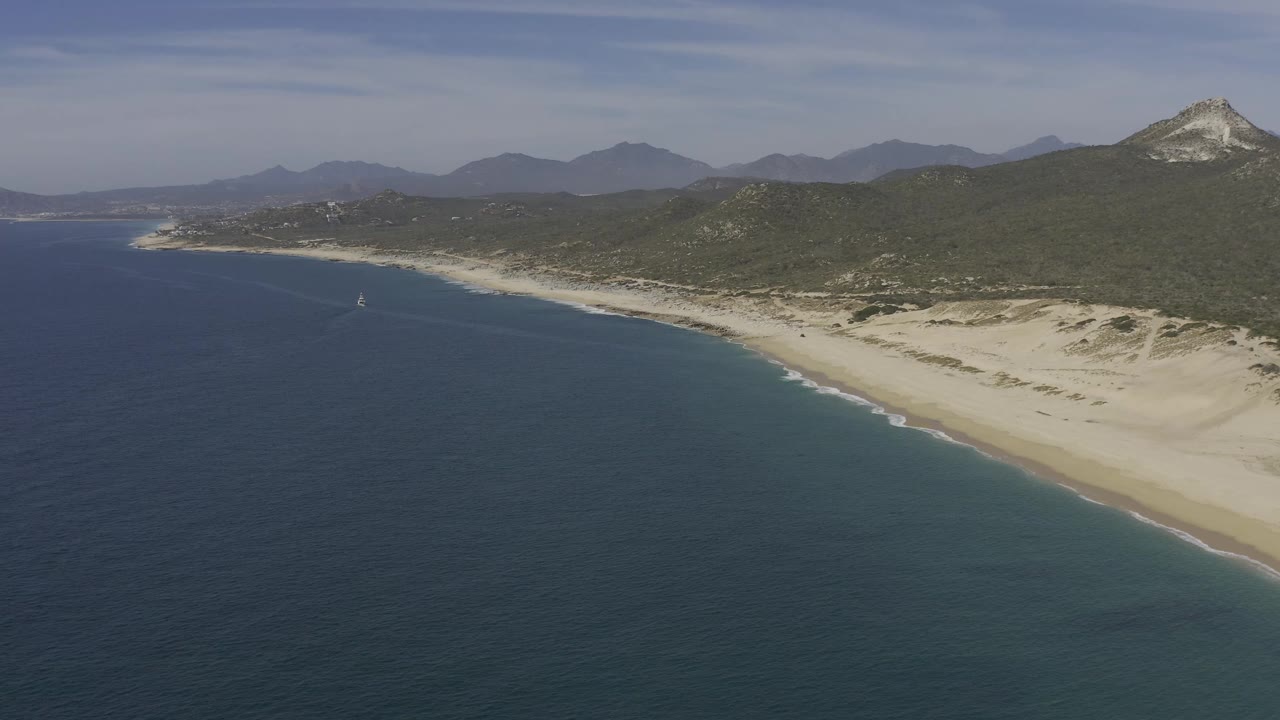 Baja California Sur aerial frame with open water filling the lower half, a long white shoreline curving across the right side, hazy mountains in the distance, and no visible people.