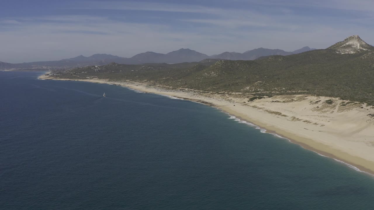 Aerial view of the Baja California Sur shore with a small sailboat on the blue water, pale beach and dunes on the right, green hills behind, and no visible people.