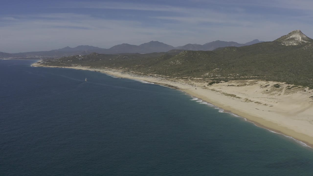Oblique aerial frame over Baja California Sur with ocean on the left, a long sandy beach and dune field on the right, hills and mountain ridges behind, and no visible people.