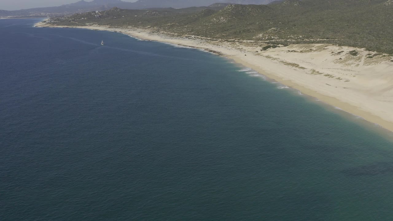 Aerial view of turquoise water beside a long sandy beach and green hills.