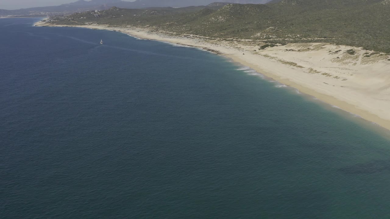 High offshore view in Baja California Sur with blue-green water filling the foreground, a diagonal sandy shoreline, low dunes, green hills, a small distant sailboat, and no visible people.