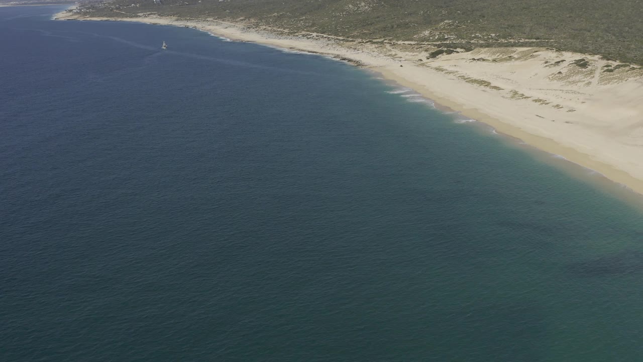 Aerial Baja California Sur coastline with turquoise ocean in front, a pale beach along the right, dunes behind the sand, distant hills, a small sailboat offshore, and no visible people.