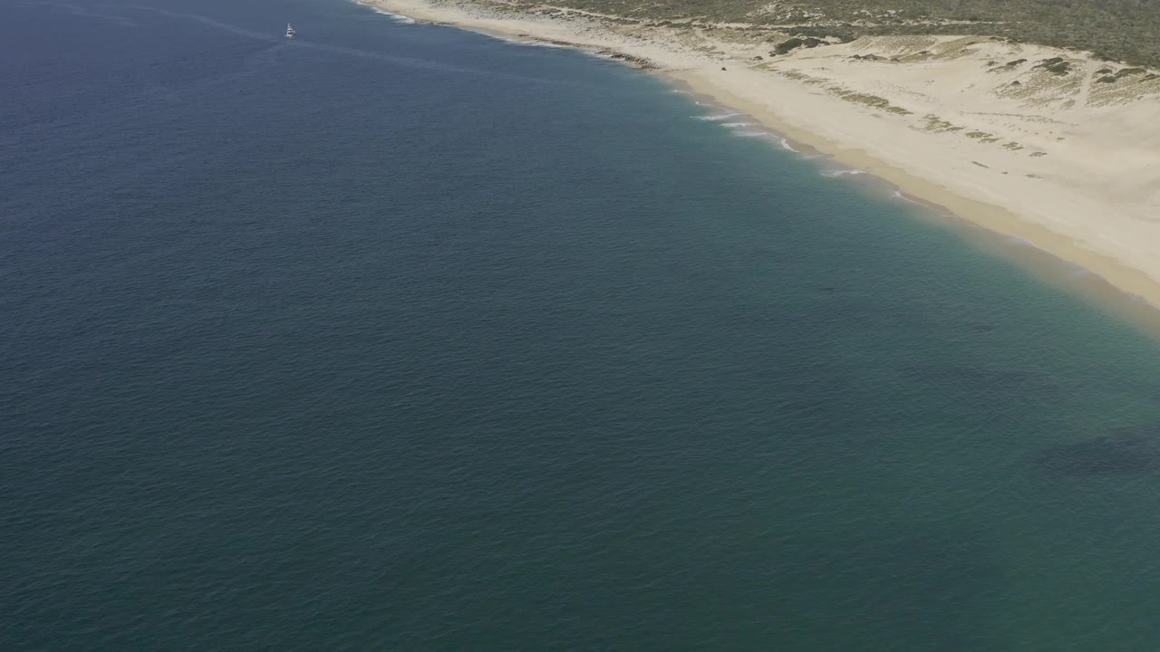 Aerial frame over open blue-green water in Baja California Sur, with the sandy beach and dune line crossing the upper right, a faint distant sailboat, and no visible people.
