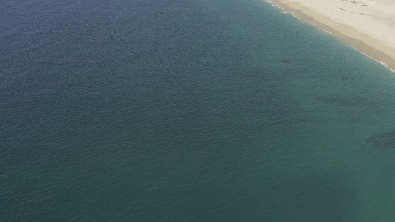 Baja California Sur aerial view with blue-green ocean dominating the frame, a thin sandy shoreline near the top, and no visible people.