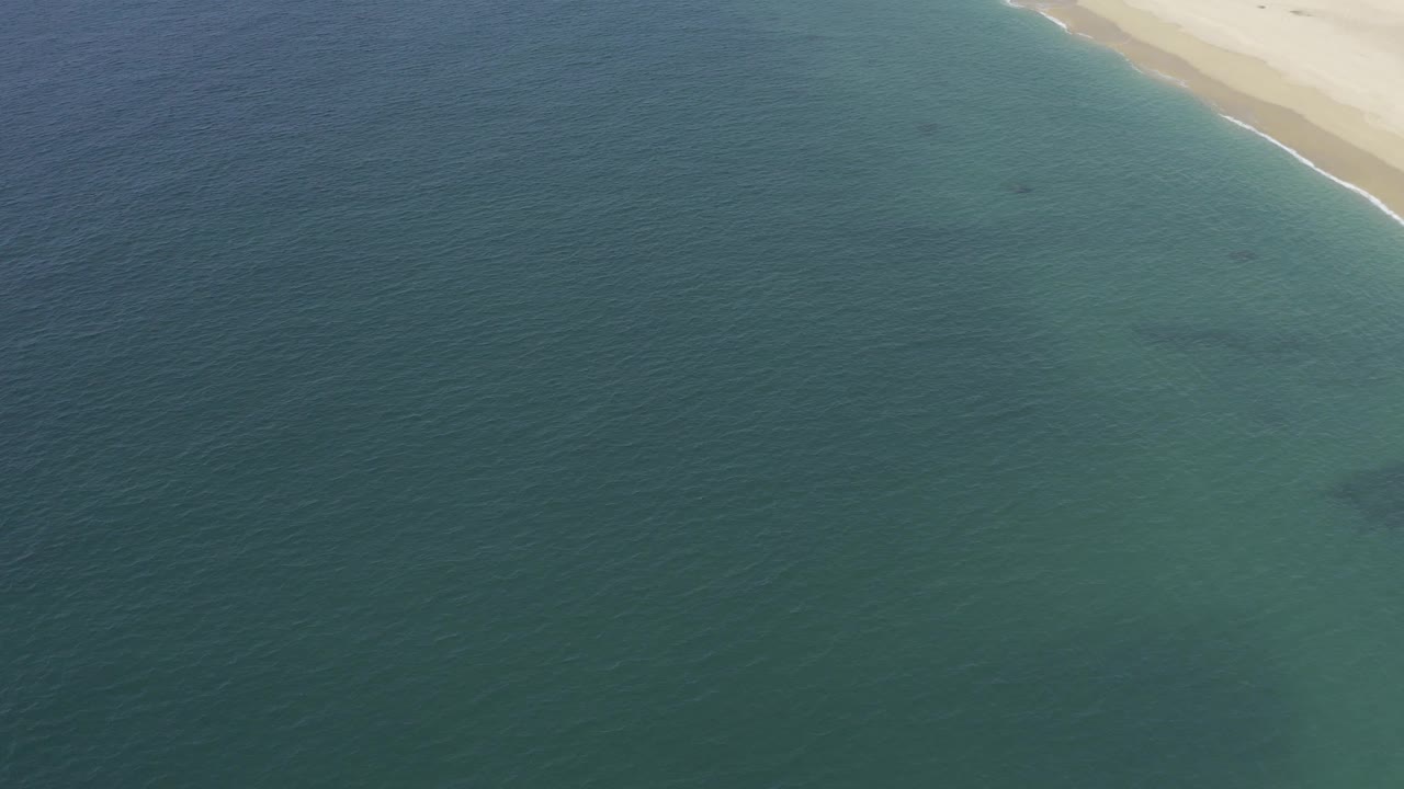 Aerial coastal frame in Baja California Sur showing clear water gradients from turquoise to deeper blue, bright sand near the top, and no visible people.