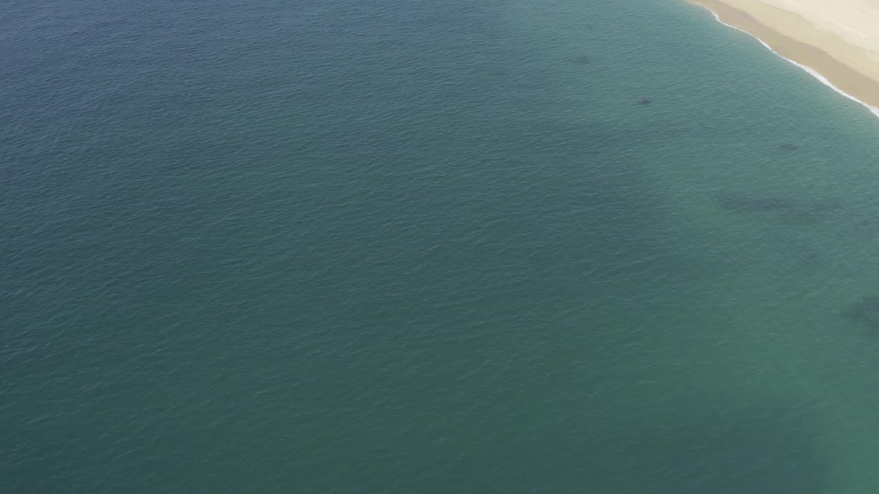 Baja California Sur shoreline from above with shallow green water in the midground, darker patches below the surface, pale sand at the top, and no visible people.