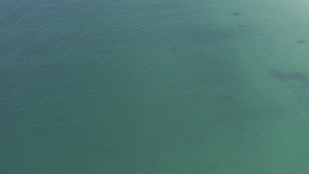 Top-down view of Baja California Sur water, with a darker gray underwater shape near center-left, teal surface color, fine ripples, and no visible people.