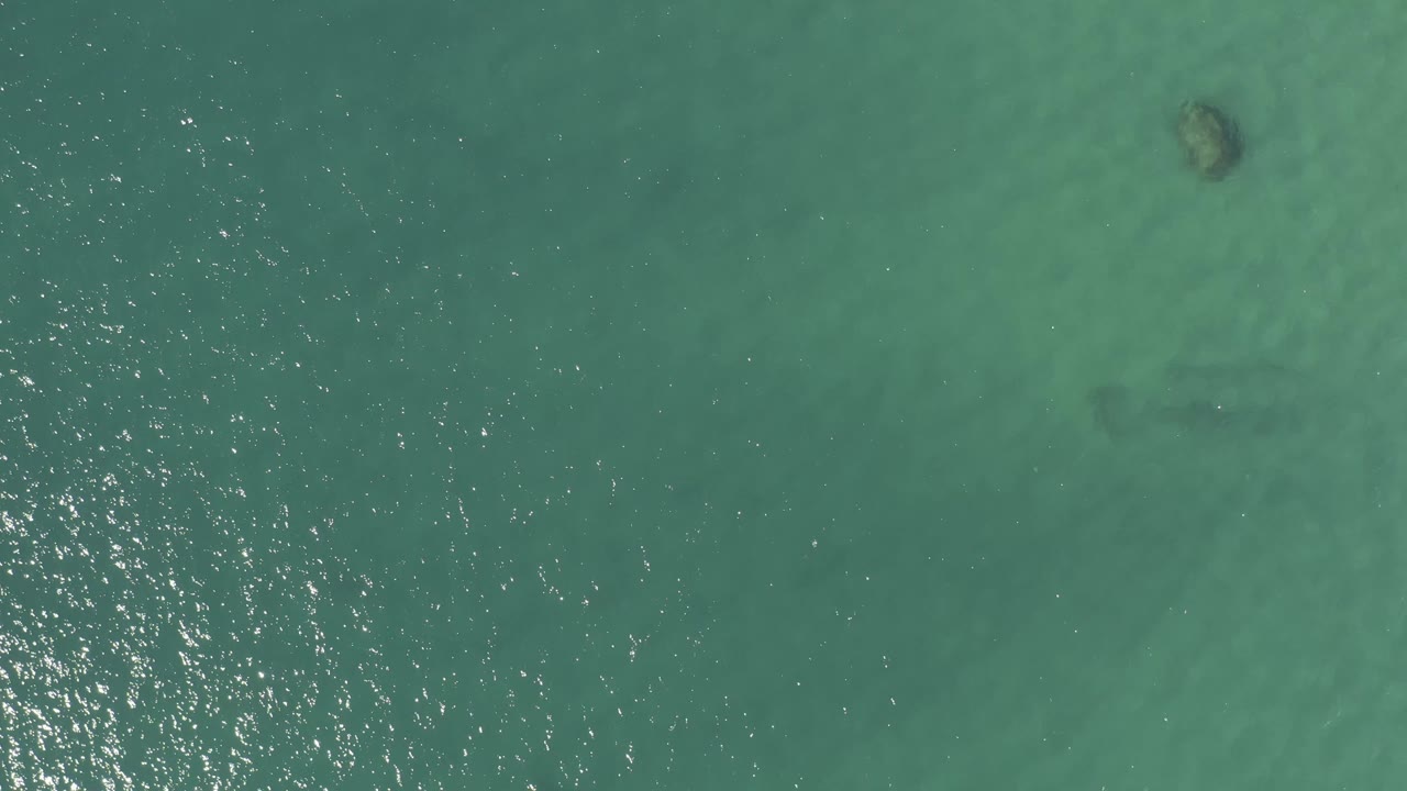 Top-down view over Baja California Sur coastal water with a bright vertical strip of sun reflections crossing green-blue ripples and faint seabed marks, no visible people.