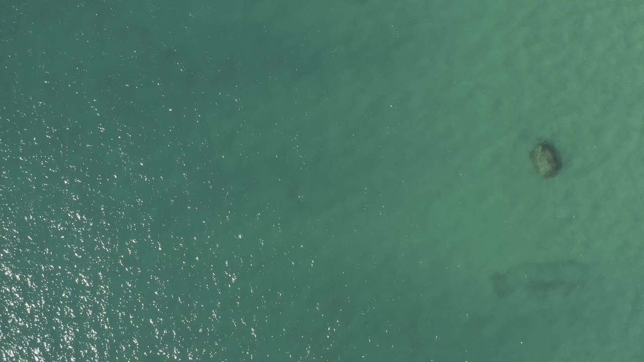 Overhead Baja California Sur water with a round darker rock or coral head near the lower center, pale sand-colored bottom, and glittering ripples along the left edge, no visible people.