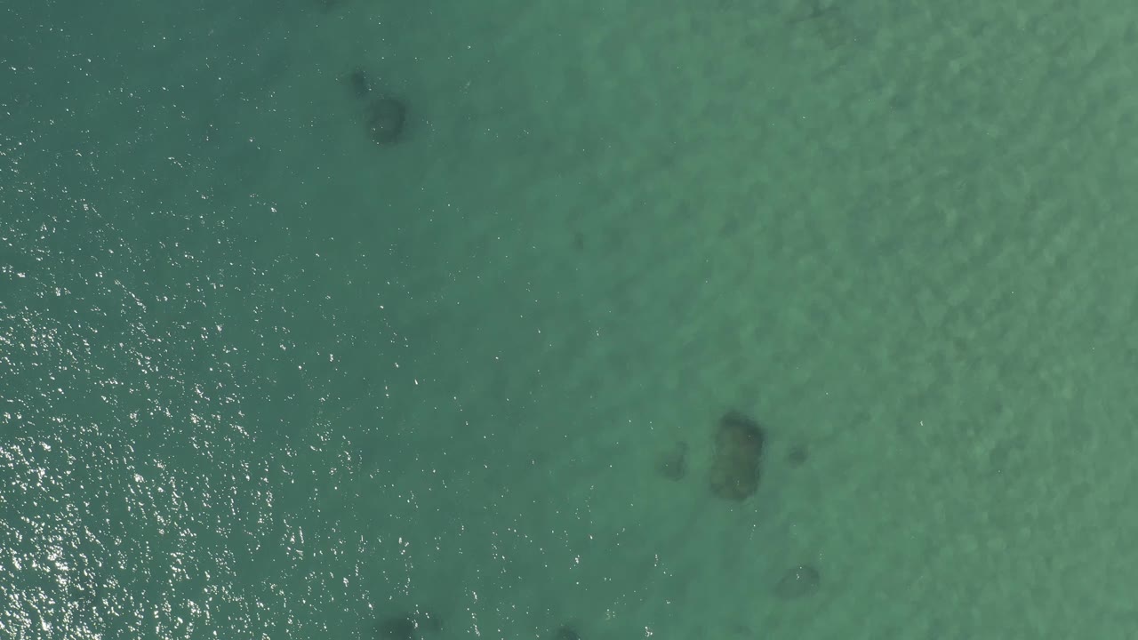 Top-down Baja California Sur water view with a bright band of reflected light through the middle, scattered dark submerged patches, and no visible people.