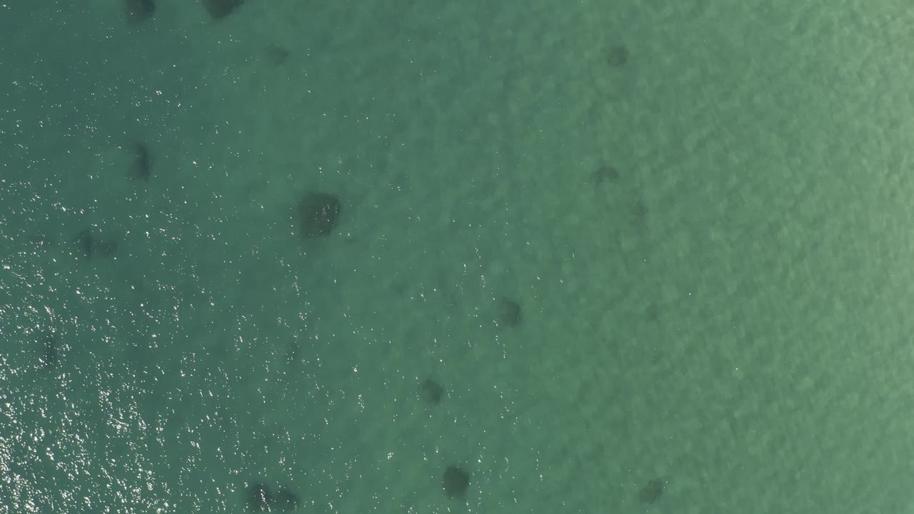 Aerial view in Baja California Sur with textured turquoise water, dark patches below the surface, bright reflected light, a small white surf burst near the right edge, and no visible people.