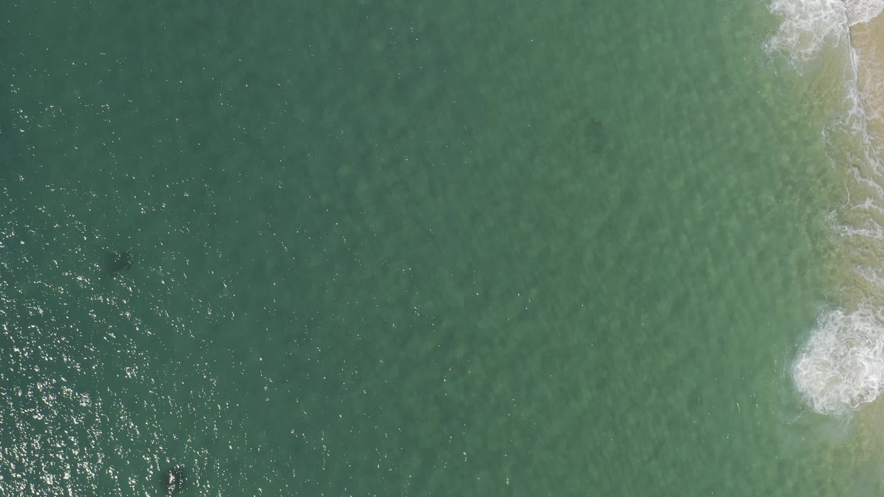 Overhead view in Baja California Sur, Mexico, with blue-green water filling the frame, a small white surf break near the right edge, and no visible people.