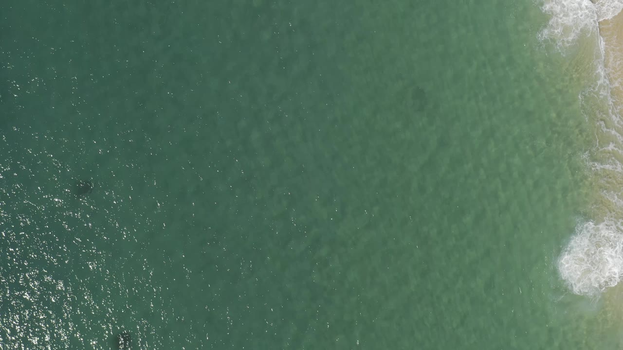 Aerial view in Baja California Sur, Mexico, showing a wider field of blue-green water with bright sun speckles, a foamy break near the right side, and no visible people.