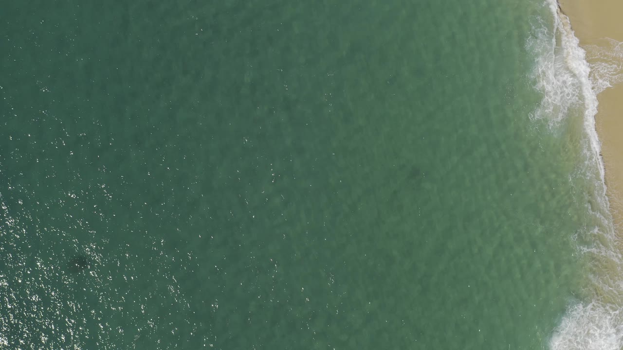 Top-down coastal view in Baja California Sur, Mexico, where blue-green water meets a pale sandy beach, with white foam curling along the right edge and no visible people.