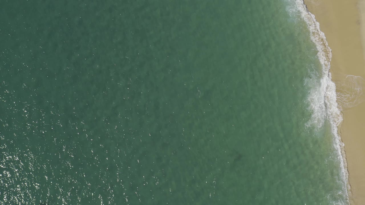 Aerial frame from Baja California Sur, Mexico, with the sandy beach pressed along the right side, blue-green water across the left, a foamy wave at midframe, and no visible people.