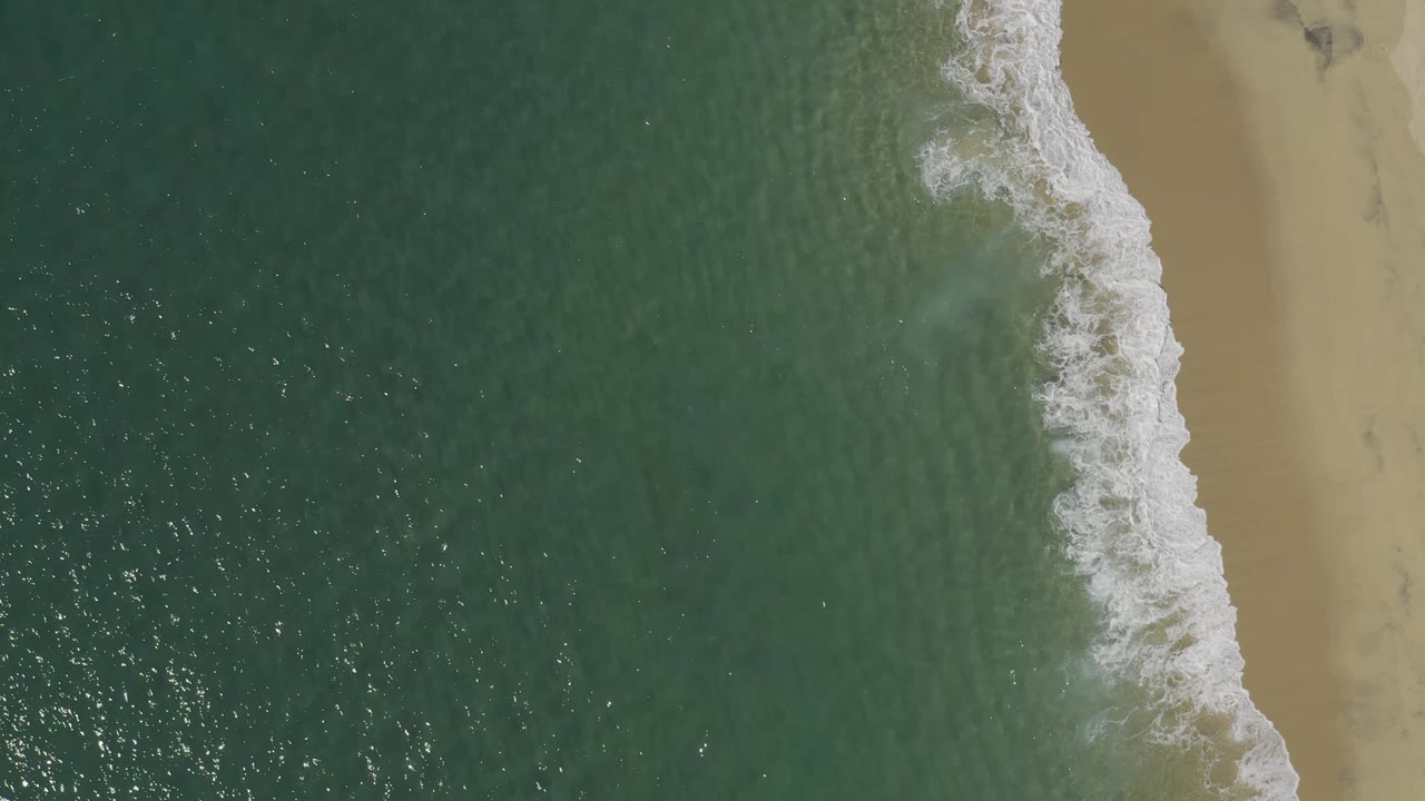 Aerial frame from Baja California Sur, Mexico, showing white wash sliding along the edge of green water and pale sand with no visible people.