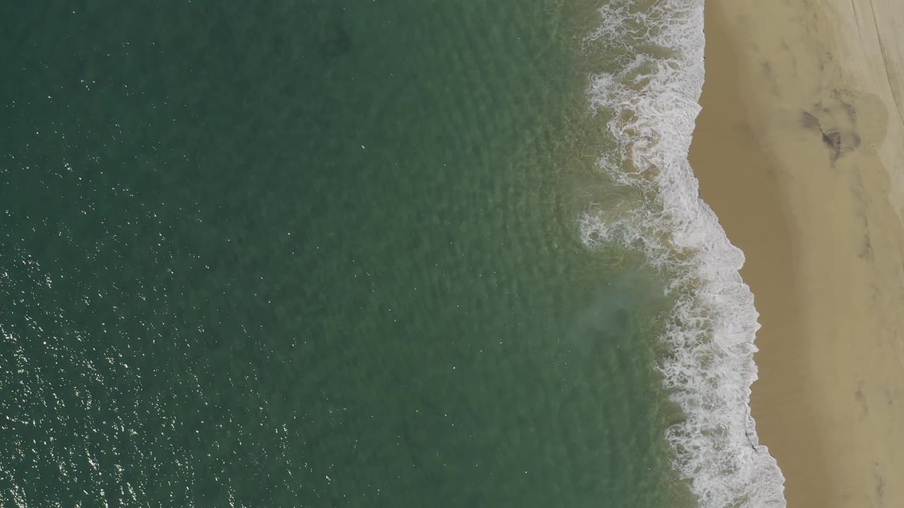 Top-down Baja California Sur, Mexico shoreline view with darker green water, bright foam near the beach edge, sandy right side, and no visible people.