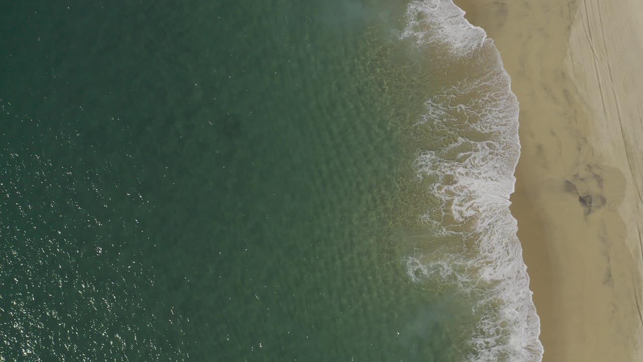 Drone perspective over Baja California Sur, Mexico, with textured green water, foamy wave wash, tan sand, faint beach tracks, and no visible people.