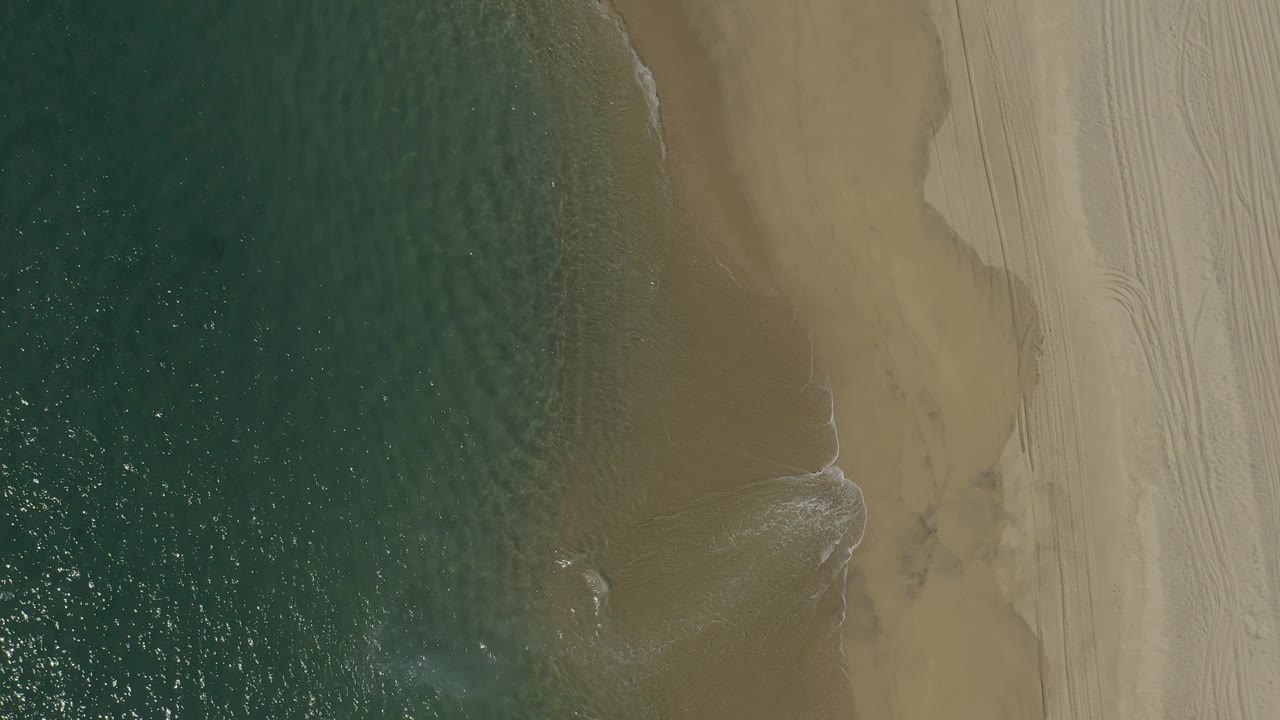 Drone view over the Baja California Sur coast with textured green water at left, a narrow foam seam at the shore, and open tan sand with no visible people.